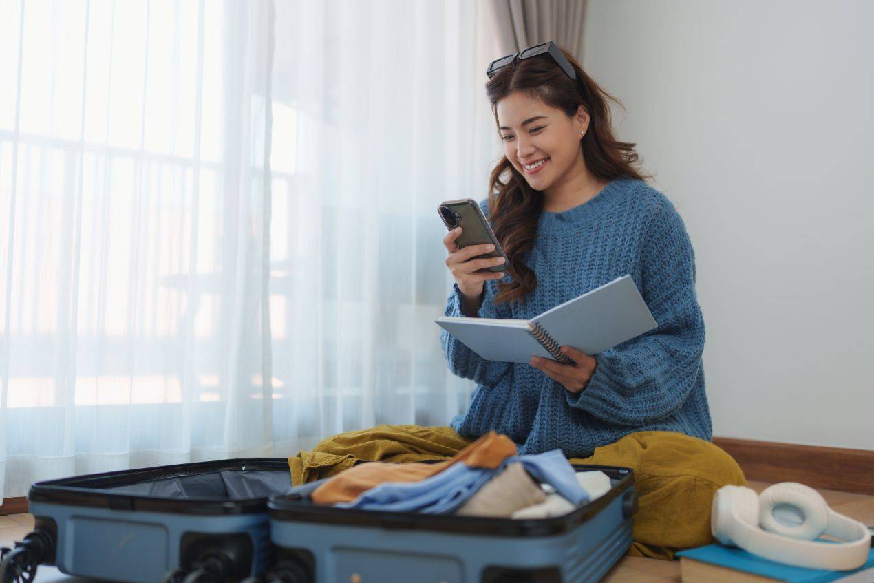 Junge Frau bereitet sich im Hotelzimmer auf ihre Reise vor, mit Smartphone, Notizbuch und offenem Koffer – Eurostecker und Reisegadgets gehören zur Ausstattung
