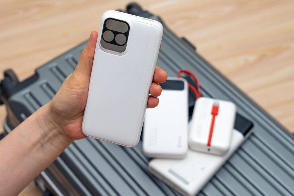 Mehrere Powerbanks liegen auf einem Reisekoffer, eine Hand hält ein Smartphone mit integrierter Akkuhülle – perfekte Ergänzung zum Eurostecker im Handgepäck