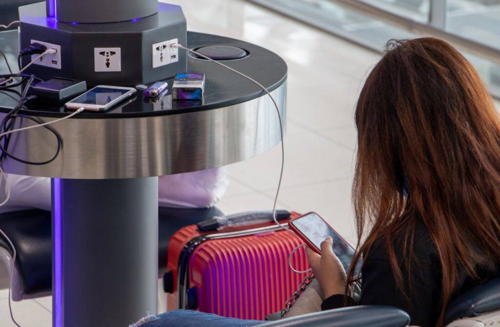 Reisende sitzt am Flughafen mit Koffer und lädt ihr Smartphone an einer öffentlichen Ladesäule mit internationalen Steckdosen – Eurostecker nicht passend