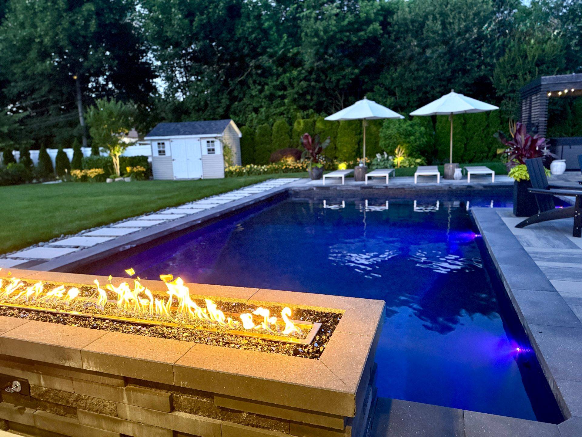 Moderner Garten mit beleuchtetem Pool und Feuerstelle bei Sonnenuntergang